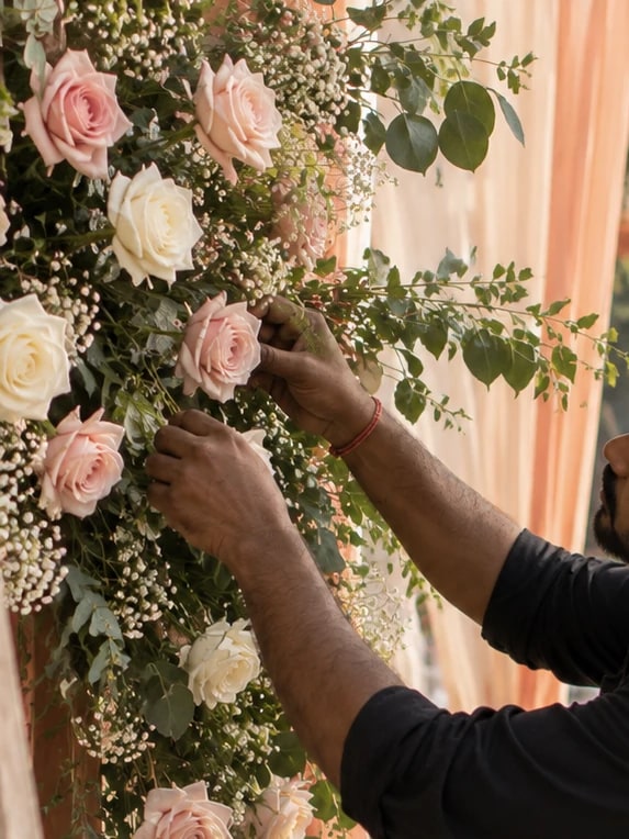Close-up of a decorator’s hands fixing blush and cream roses, gypsophila and eucalyptus onto a floral backdrop at a wedding setup